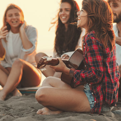 Millennial woman playing her guitar, surrounded by a group of friends on the beach
